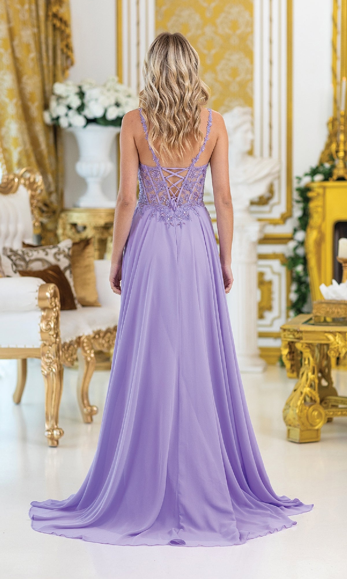 A woman with wavy blonde hair stands in an ornate room, wearing the Dancing Queen 4531 Lace-Corset Long Prom Dress in lavender, featuring elegant embroidery and intricate lace-up back detailing.